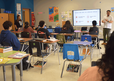 Classroom setting with students listening to a teacher presenting on a screen