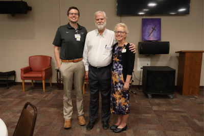 Three people smiling in a conference room setting with chairs and a cross painting in the background