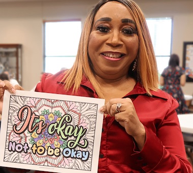 Smiling person holds sign reading Its okay not to be okay with colorful floral design in office setting